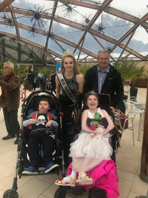 A man and woman stand behind a boy and girl in wheelchairs. The woman is dressed in a pageantry outfit and a tiara. The boy is dressed as a pirate and the girl as a princess.