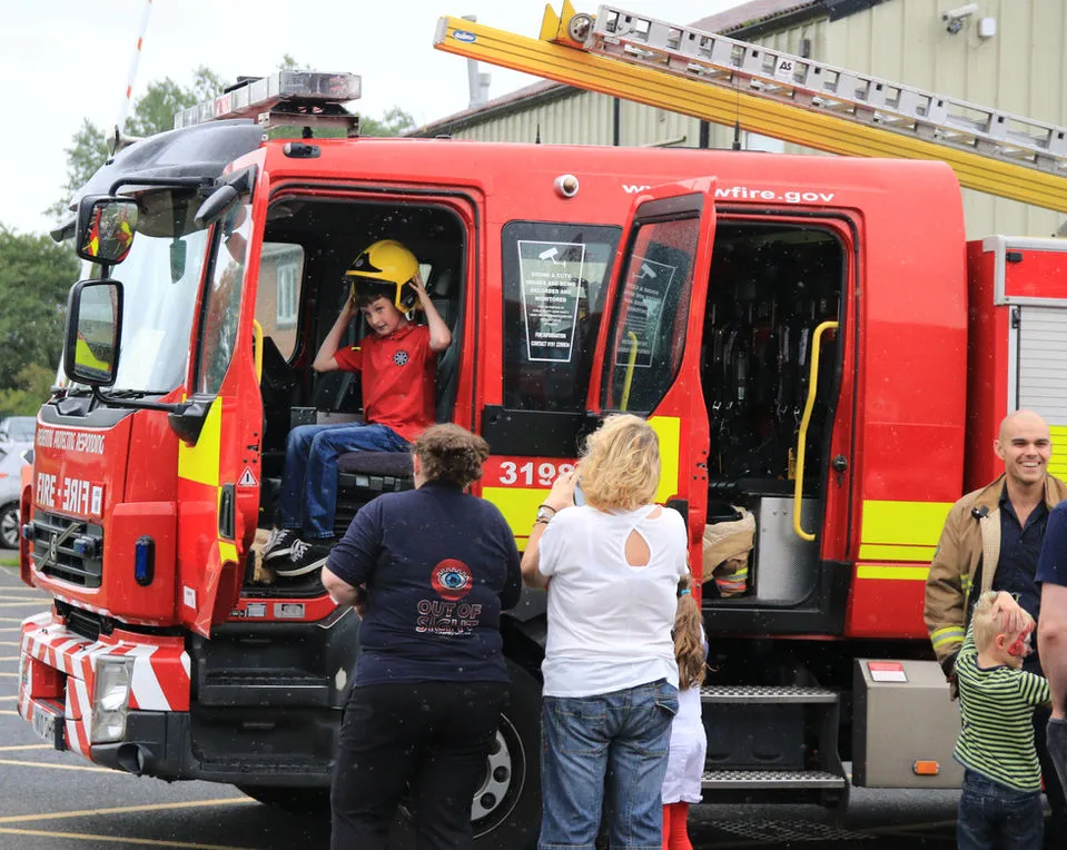 Two women are looking at a boy sitting in the passenger's seat of a fire enginge truck. He is trying on a fireman's helmet.
